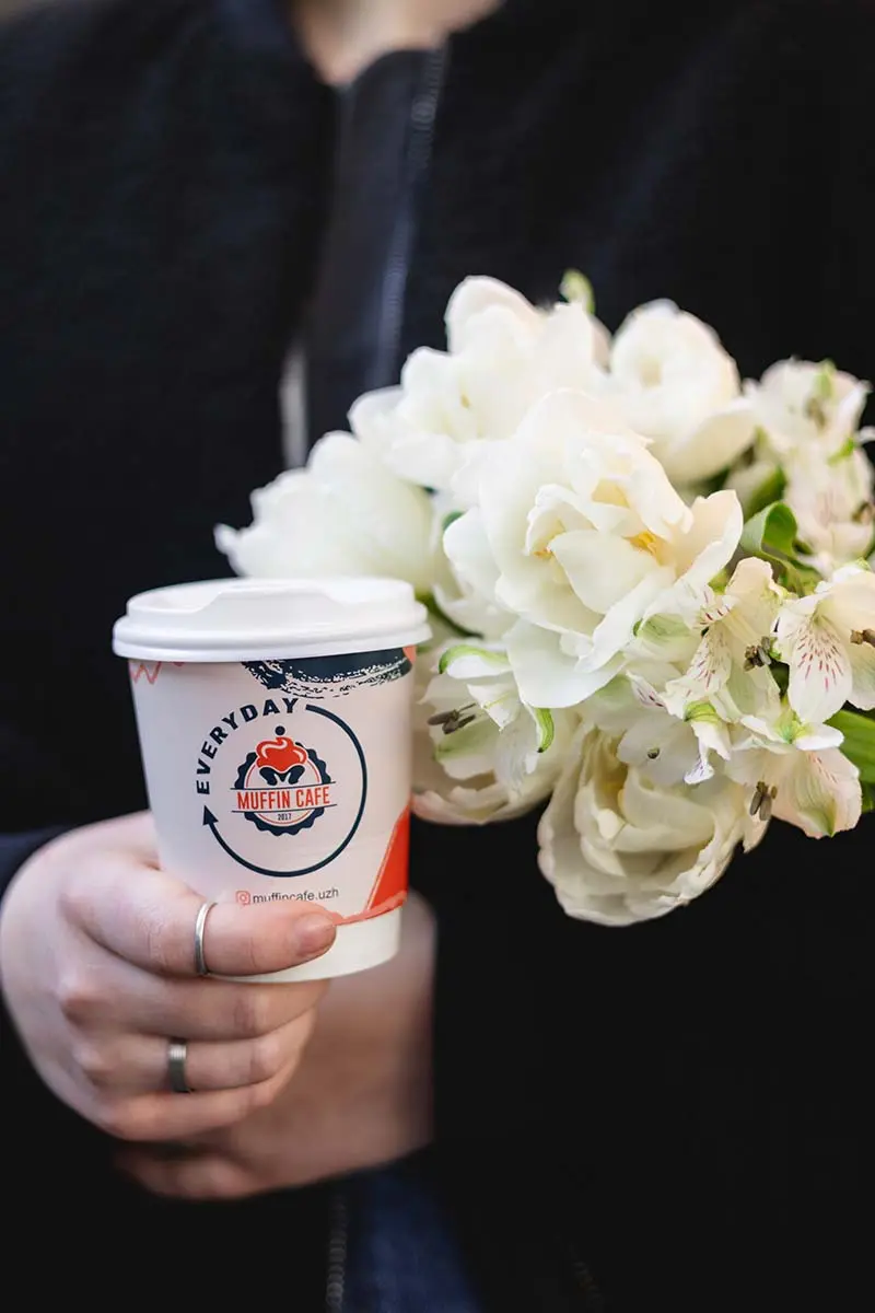 Дівчина тримає каву to-go з логотипом Muffin Cafe та букет білих квітів.